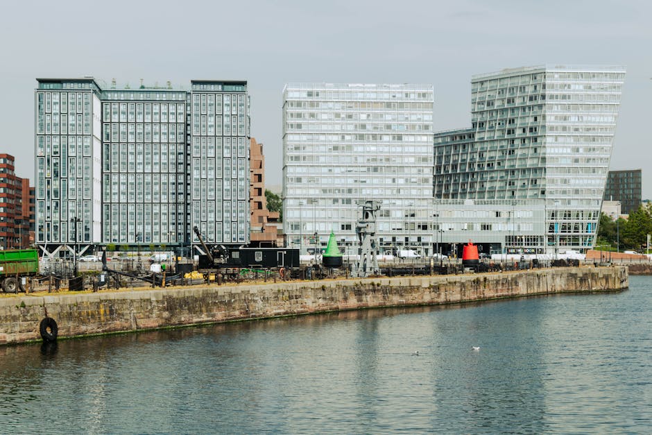 A high-angle view of a modern urban canal surrounded by tall commercial office buildings made of glass, steel, and stone on both sides. The water reflects the buildings and the partly cloudy sky overh